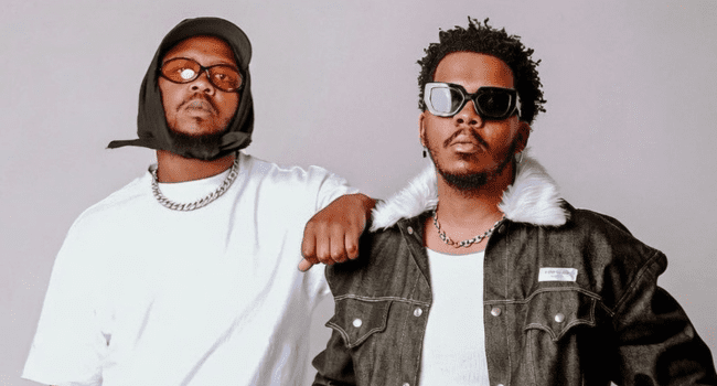 Two men pose together; one sports a white shirt, black hat, and sunglasses, while the other—channelling the style of Stino Le Thwenny—wears a black jacket with fur collar and dark sunglasses.