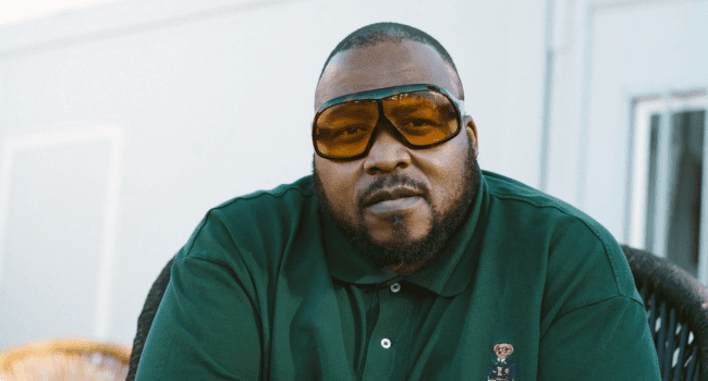 A man with a beard wearing large amber-tinted glasses and a green polo shirt sits outdoors, looking directly at the camera, channeling the cool confidence of rap legends like Stogie T.