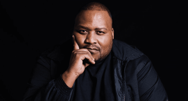 A man wearing a black jacket and black shirt sits against a dark background with his hand resting on his chin, looking at the camera—evoking the contemplative style often seen in SA Hip-Hop icons like Stogie T.