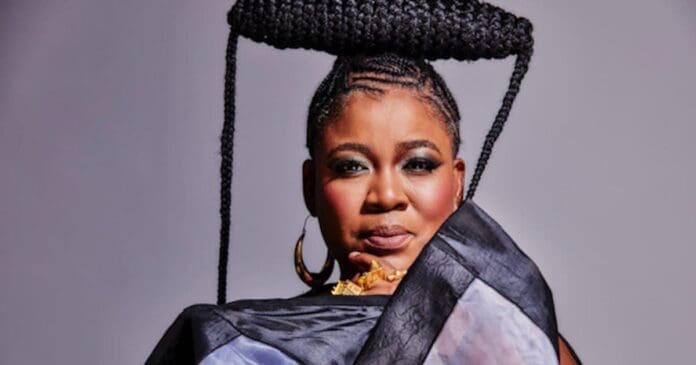 A woman with intricate braided hair styled into a large, horizontal shape poses in a dark, patterned outfit against a plain backdrop, evoking the iconic style of Thandiswa Mazwai and celebrating rich history.