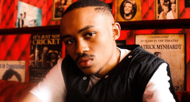 A young man in a black vest sits indoors against a red wall decorated with vintage posters, including one featuring PRESS HASH: VOLUME ONE.