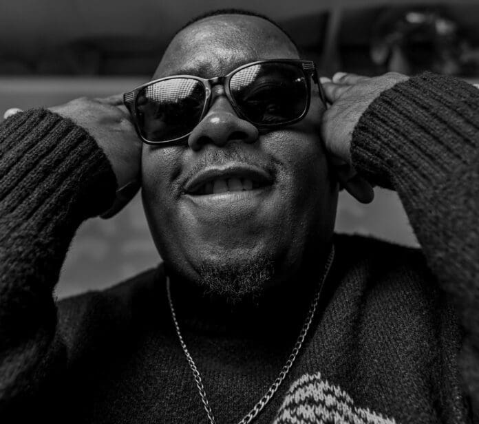 A man wearing dark sunglasses and a sweater holds his hands to the sides of his head and smiles, photographed in black and white—capturing the cool energy of Amapiano.