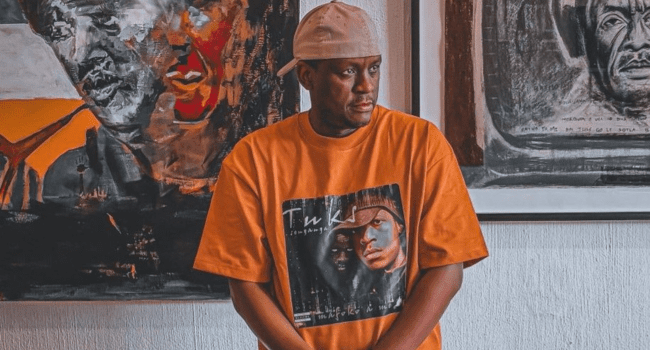A man wearing an orange graphic T-shirt and a backward beige cap stands in front of colorful abstract art paintings, channeling the energy of Tuks Senganga’s Balance as he teases tracks from his upcoming EP.