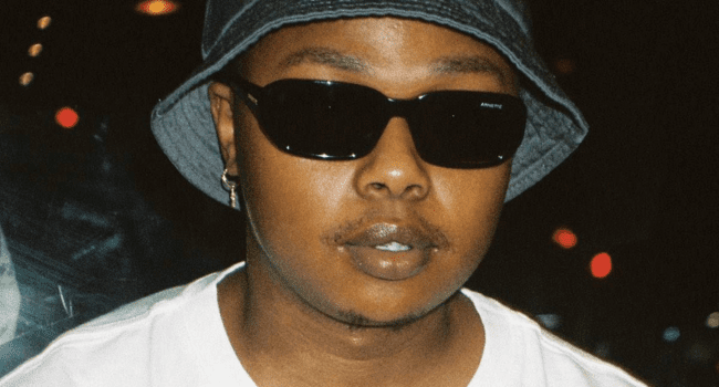 A person wearing a bucket hat, dark sunglasses, a white shirt, and an earring stands indoors with a neutral expression, channeling the cool style often seen on artists like Stogie T.