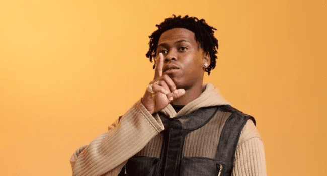 A person stands against a plain orange background, holding a finger to their lips in a gesture signaling silence—channeling the cool confidence of US Rapper Loe Shimmy.