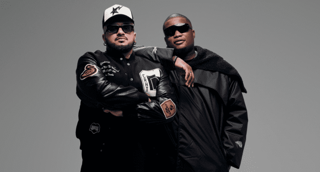 Two men wearing black jackets and sunglasses pose together against a plain gray background, embodying the Duo Venom vibe. One has his arms crossed while the other rests his arm on his companion's shoulder.