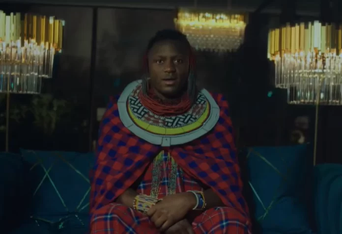 A person wearing traditional Maasai attire sits on a blue couch between two chandeliers in a dimly lit room, evoking the vibrant style seen in Burna Boy's new music video.