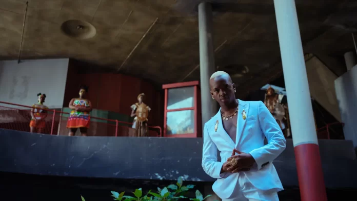 A man in a white suit stands in the foreground, while four African artists in traditional attire stand on a balcony in the background of a modern concrete building.