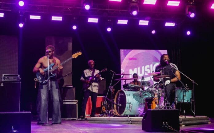A band delivers a dynamic production on stage under purple lights at Music City Central; the bassist sings into a microphone while the guitarist and drummer play their instruments.