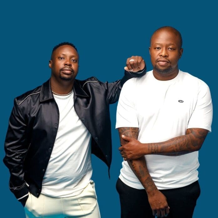 Two men stand side by side against a blue background; one wears a black jacket and white pants, while the other wears a white t-shirt and shows tattoos. Their style channels the vibrant energy of Sphectacula & DJ Naves.