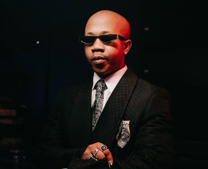 A man wearing a black suit, patterned tie, and sunglasses stands with folded arms against a dark background, channeling the cool confidence of Nasty C.