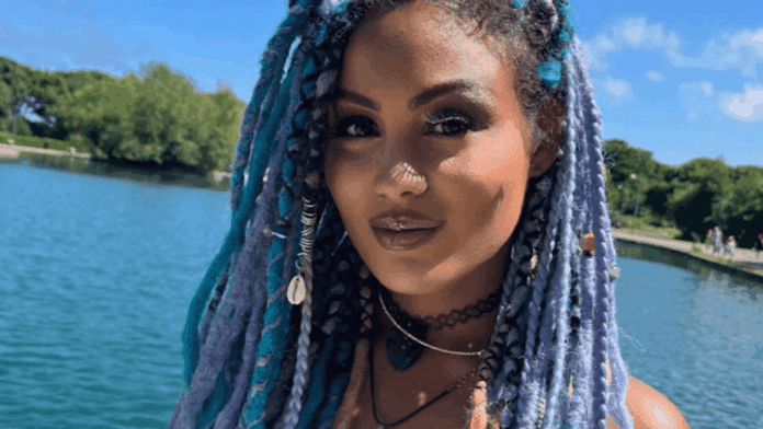 A woman with long, blue and purple braided hair stands by a body of water on a sunny day, exuding Afrobeats vibes with her black choker and metallic lipstick. Trees and blue sky provide a serene Melody in the background.
