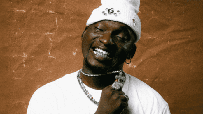 A person in a white beanie and shirt holds a pendant on a silver chain, smiling with eyes closed against a brown background, celebrating Amaed's new single "Ayanmo.