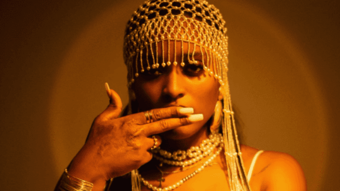 A person wearing a beaded headpiece and pearl jewelry holds their hand near their mouth, with warm yellow lighting in the background—an image inspired by Nino Mayanna’s new single "Aje.