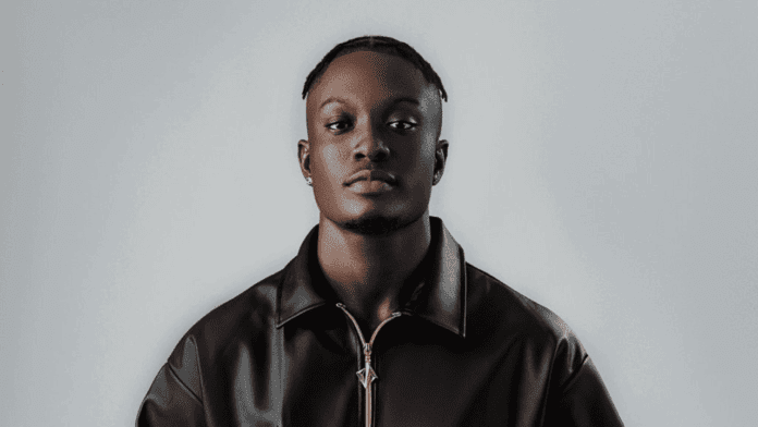 A man with short braids, wearing a black leather jacket and earrings, stands against a plain light gray background, looking directly at the camera—MC Shocker, known for "Was Once In Love," teases his new EP.