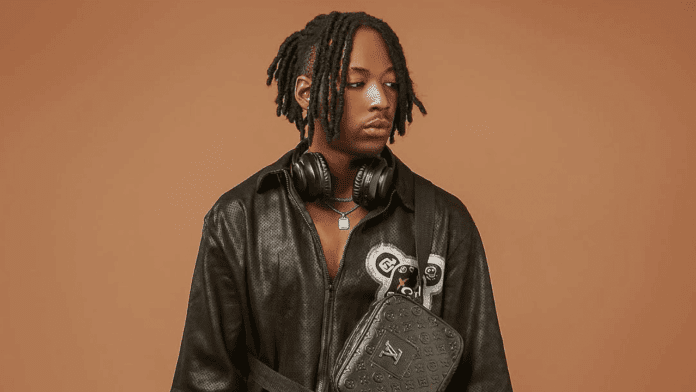 A person with short dreadlocks wears a black leather outfit, headphones around the neck, and carries a black Louis Vuitton bag, standing against a plain brown background, ready to vibe to Lil Tizzy’s new single "No Be Today.