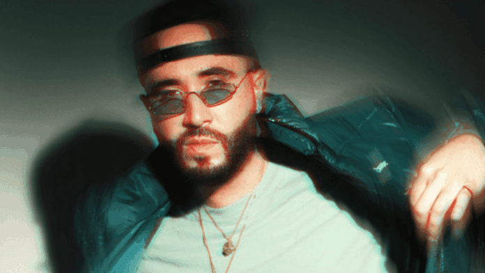A man with a beard wearing round sunglasses, a backward cap, and layered necklaces poses in a puffer jacket against a plain background with a blur effect, channeling Afrobeats style for his new single.