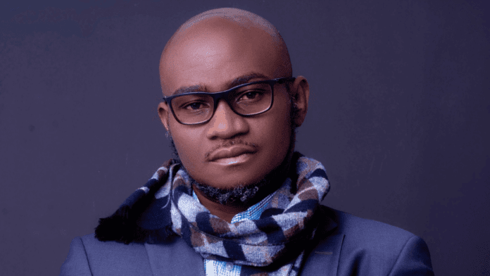 A man with glasses, a patterned scarf, and a suit jacket poses against a plain dark background, evoking reflections on Time and Chance.