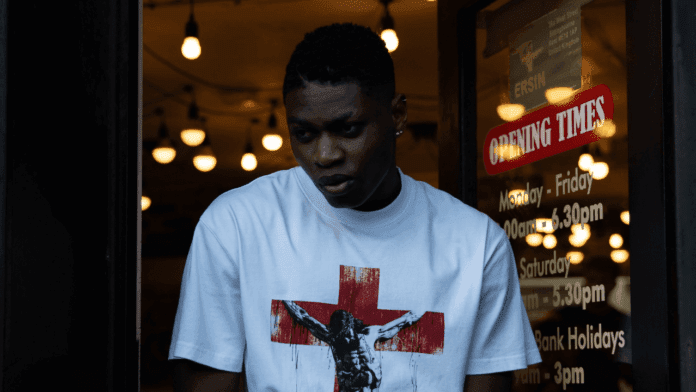 A person in a white T-shirt with a red cross and Jesus graphic stands near a door displaying shop opening times, as warm lights glow inside—giving the scene a dream-like feel. The word "JAREH" is subtly printed on the shirt’s sleeve.