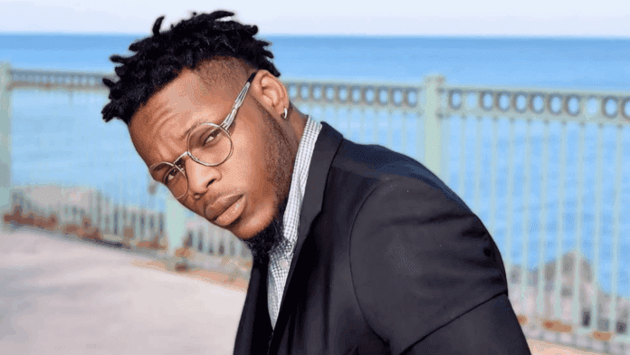 Man in a black suit and glasses leans slightly forward, posing outdoors near a metal fence with water and sky in the background, radiating Jessenation vibes—a style as striking as an Afrobeats love song.