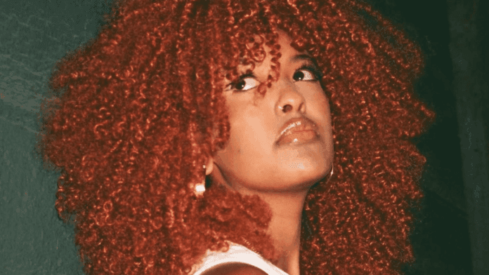 A person with voluminous, curly red hair looks off to the side against a dark green background, wearing a white top and earrings—channeling Aleeta's spirited style with an effortless grace.