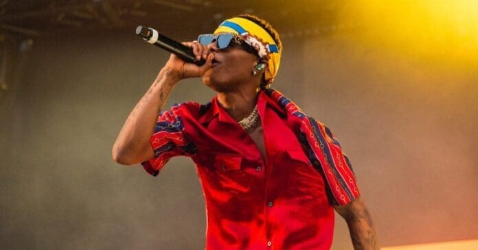A performer wearing a red shirt, sunglasses, and a colorful headband sings into a microphone on stage under yellow lighting at Afro Nation 2026.