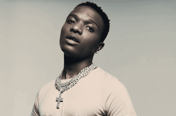 A young man with short hair wearing a beige t-shirt and a thick silver chain necklace with a cross pendant, reminiscent of Wizkid's style, stands against a plain background, focusing on the important things in life.