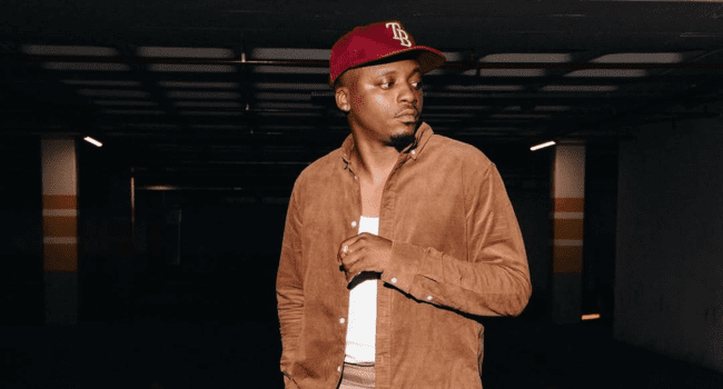 A man wearing a brown jacket, white shirt, and red baseball cap stands in a dimly lit indoor parking area, looking to his left—as if anticipating a high-octane clash reminiscent of a Cricket T20 showdown.