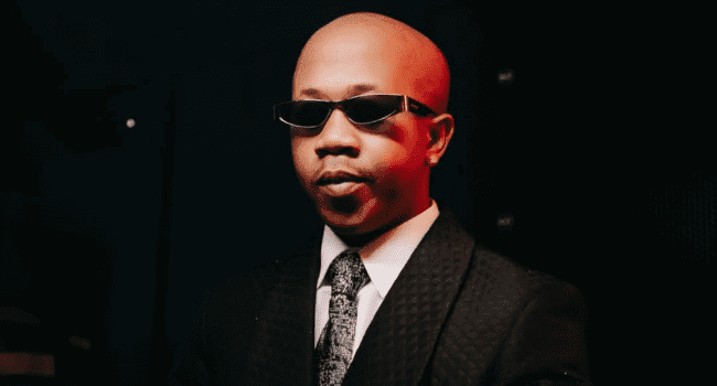 A man in a black suit, patterned tie, and sunglasses stands against a dark background with red lighting, channeling La Vida Loca vibes reminiscent of Nasty C’s bold style.