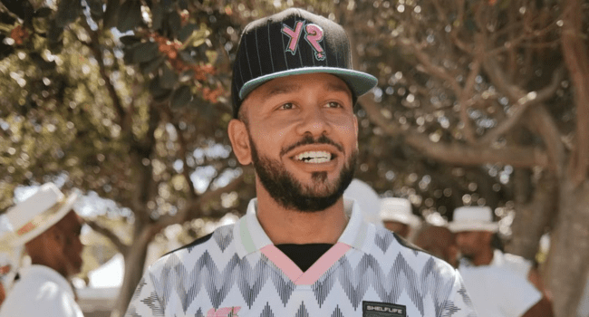 A man with a beard and mustache wearing a black cap and patterned shirt stands outdoors, with trees and people in hats in the background—reminiscent of YoungstaCPT at the peak of his music career.
