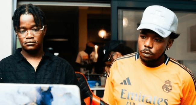 Two men stand side by side indoors, one in glasses and a black shirt, the other in a light cap and yellow "Emirates" jersey, both gazing off-camera—capturing the vibe of SA Hip-Hop producers like Mashbeatz and Zoocci Coke Dope.