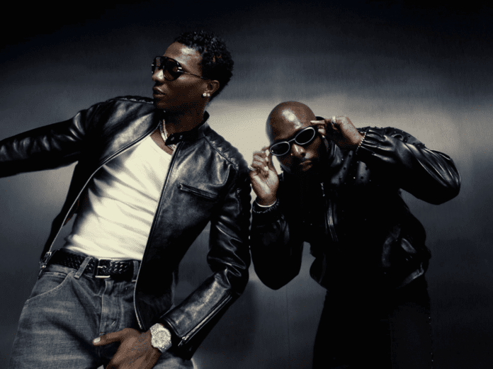 Two men in black leather jackets and sunglasses pose against a dark background; channeling Jogodo energy, one leans left with a hand out, while the other adjusts his glasses.