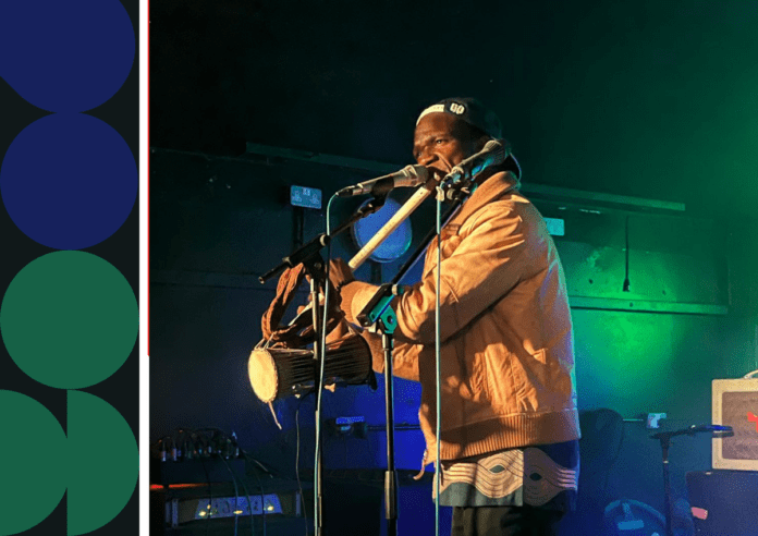 A musician wearing a tan jacket and cap plays a traditional drum and sings into two microphones on the darkly lit stage of Attic Bar.