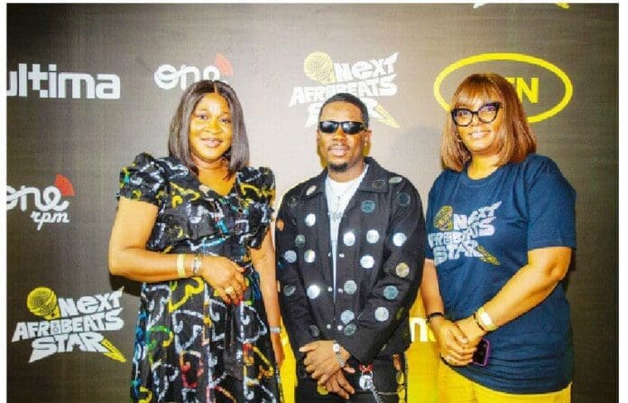 Three people stand in front of an MTN Next Afrobeats Star backdrop. The man in the middle, Ayo Benzi, sports sunglasses and a black outfit, while the two women beside him wear vibrant, patterned ensembles reflecting the Afrobeats spirit.