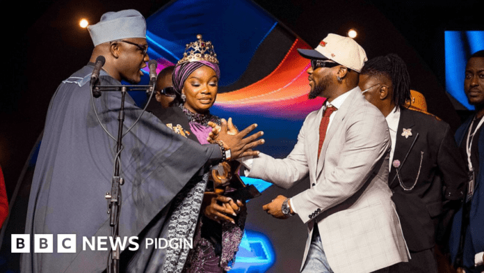 A woman wearing a crown and sash accepts the Artiste of the Year award on stage from two men in suits at the Ghana Music Awards 2025, as several other people look on. BBC News Pidgin logo appears at the bottom left.