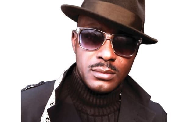 A person channeling Afrobeats flair, wearing a black hat, dark sunglasses, a black turtleneck, and a dark coat, looks directly at the camera against a plain white background.