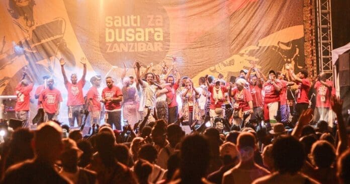 A large group of performers stands on stage waving to a crowd at the Sauti za Busara African music festival in Stone Town, Zanzibar.