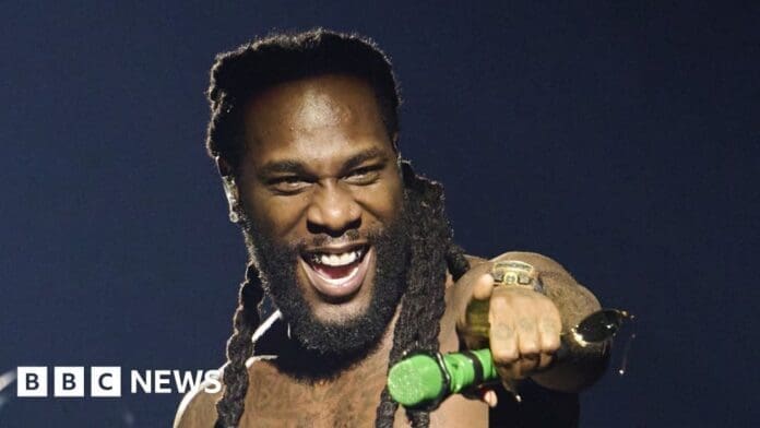 A man with long dreadlocks smiles while performing on stage, holding a green microphone and pointing forward. Nigerian stars like Burna Boy often light up such moments. BBC News logo appears in the corner.