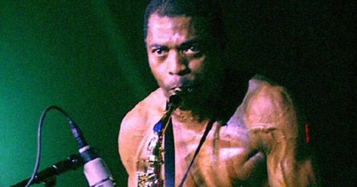 A shirtless man, reminiscent of African artist Fela Kuti, plays a saxophone on stage in front of a microphone with a dark green background.