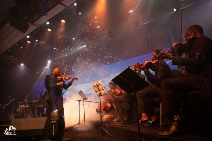 An Afrochestra dressed in black performs on stage with violins and cellos, surrounded by stage lights, smoke, and bubble effects.