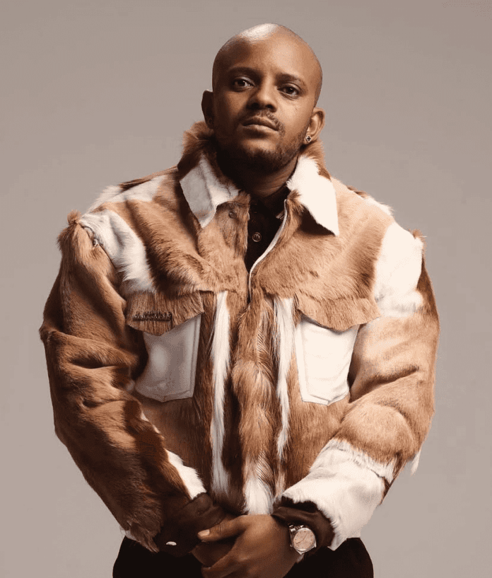 Kabza De Small stands facing the camera, wearing a brown and white fur jacket over a black shirt, with a neutral background.