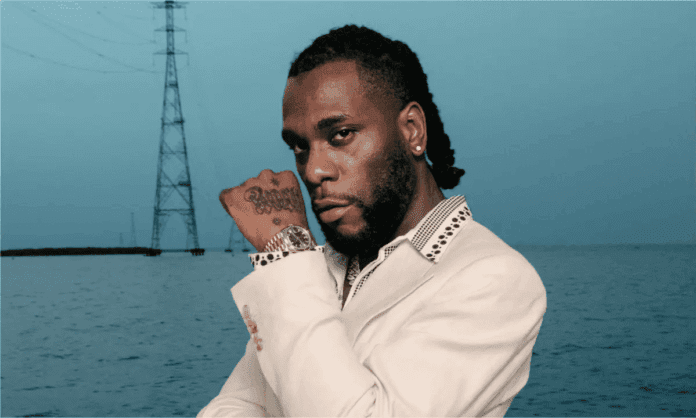 A man in a light-colored suit poses by a body of water with a power line tower in the background. Channeling Burna Boy, the renowned African artist known for his highest-grossing tour, he shows a tattooed hand and wears a watch.