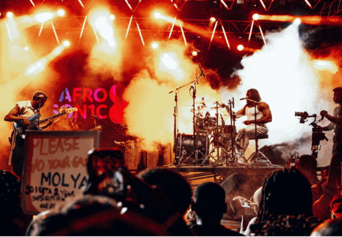 Musicians perform on a brightly lit stage at a sold-out show with "AFRO" visible in the background, as AFROSIN1C storms Accra; audience members and a sign appear in the foreground.