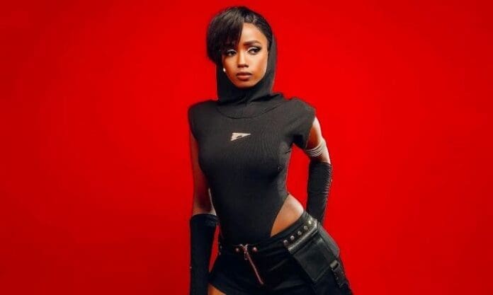 Abigail Chams, dressed in a black sleeveless hooded outfit, poses against a solid red background with one hand on her hip and an intense expression, embodying the bold energy of her "Your Loss" single.