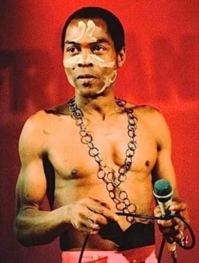 A shirtless African artist with facial paint and a chain necklace holds a microphone, standing against a red background—a powerful homage to Fela Kuti.