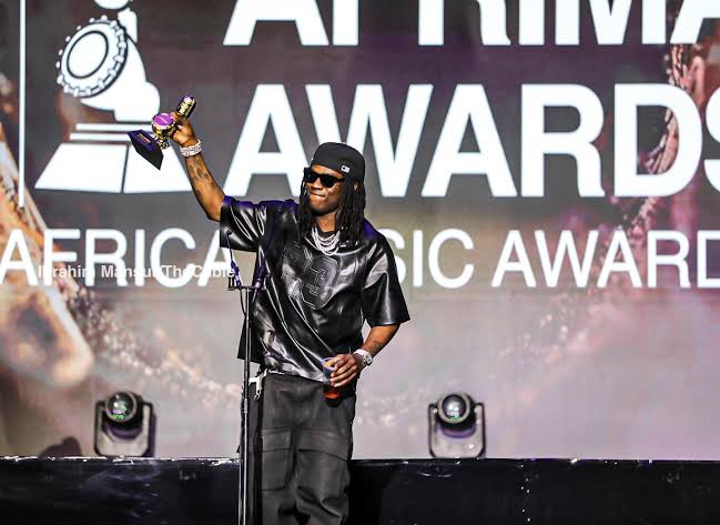 A musician in black attire holds up a trophy on stage at the AFRIMA Awards, with large event signage visible behind him—celebrating global talents like Rema and elevating the Voice of Nigeria on an international platform.
