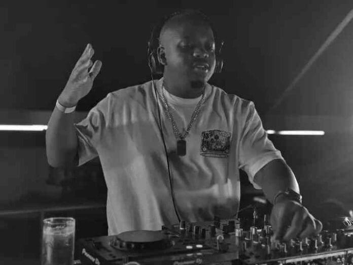 A DJ wearing headphones stands behind a mixer with one hand raised and the other on the controls; a drink is on the table nearby, spinning tracks inspired by the Spirit Fest Amapiano Mix.