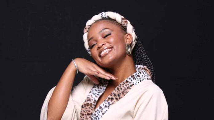 A woman, embodying Afropop vibes, wears a leopard-print collar robe and a braided headband, smiling with her eyes closed as she tilts her head and raises one hand under her chin against a black background.