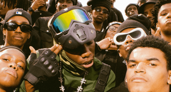 A group of young men wearing sunglasses, hats, and streetwear pose closely together, with one person in the center wearing goggles and a face mask, capturing the edgy vibe of KindlyNxsh's latest KAPTEIN music video.