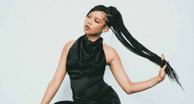 Aweh! Lordkez sits against a white background in a black dress, holding the end of her long braided ponytail with one hand—it's Perfect Timing for an iconic look.